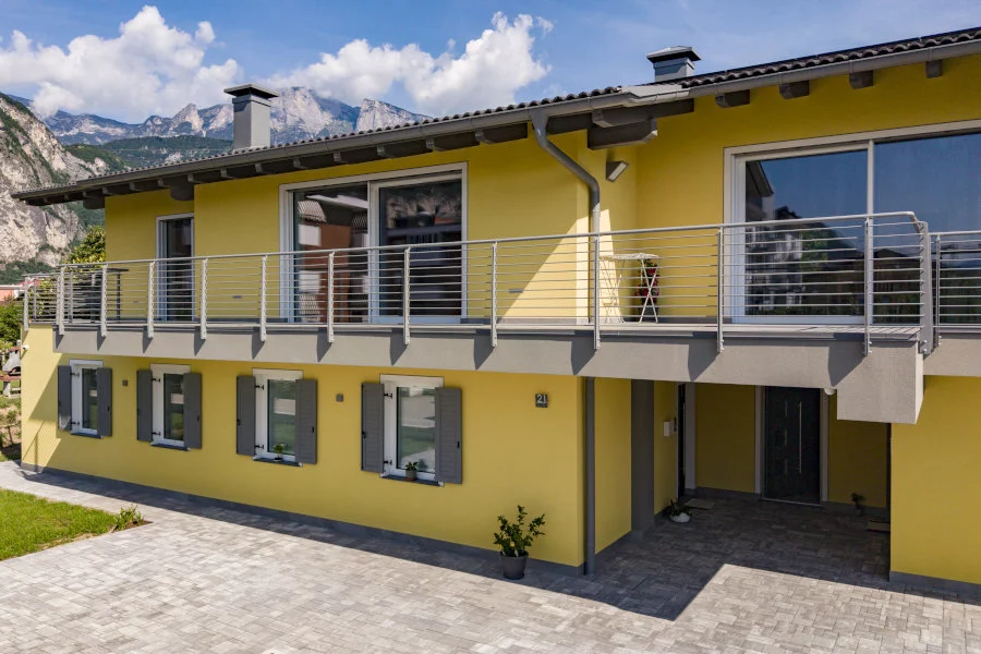 copert - Girardi Luca e Mirco s.r.l. Una casa gialla a due piani con balcone e finestre dotate di persiane grigie è visibile di fronte a unarea pavimentata, con montagne imponenti sullo sfondo sotto un cielo azzurro.