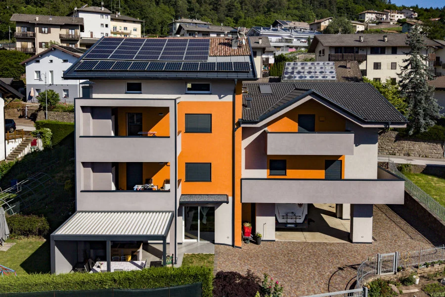 copertclasseA - Girardi Luca e Mirco s.r.l. Un moderno edificio residenziale, dalle pareti arancioni e grigie, mostra pannelli solari sul tetto, balconi e un parcheggio coperto, immerso in un contesto collinare con altre case e vegetazione.